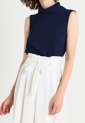 Haut sans manches bleu marine à col montant, associé à un pantalon plissé blanc taille haute avec un nœud à l'avant.