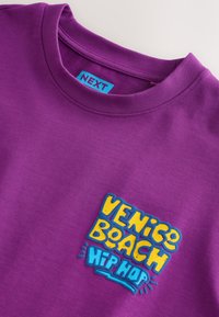 T-shirt violet à col rond avec patch de texte coloré indiquant "Venice Beach Hip Hop" et une étiquette bleue visible avec le mot "NEXT" à l'intérieur du col.