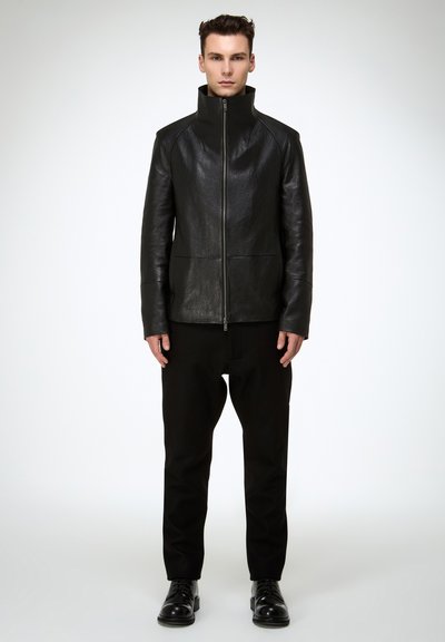 Schwarze Lederjacke mit hohem Kragen, Frontreißverschluss und zwei vertikalen Taschen. Kombiniert mit schwarzer Hose und Schnürstiefeln.