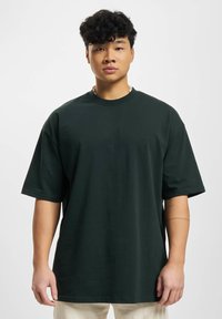 Zelené oversized bavlněné tričko s krátkými rukávy, žebrovaným kulatým výstřihem, hladkou texturou a minimálním brandingem, nošené osobou na jednobarevném pozadí.