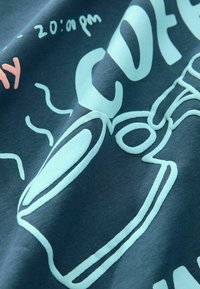 Bavlněné tričko tmavě modrozelené barvy s grafickým potiskem v světle modré a růžové barvě, zobrazující konvici na kávu a text. Hladká textura, ležérní design.