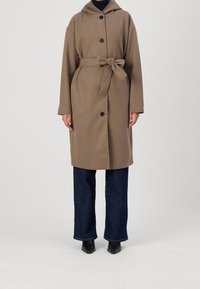 Manteau long marron avec capuche, boutons sur le devant et ceinture nouée. Caractérisé par des manches larges, il est porté avec un jean évasé bleu foncé et des bottes noires.