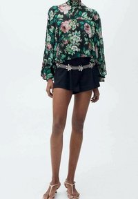 Blouse florale noire avec des fleurs vertes et roses, manches fluides, assortie à un short noir taille haute et une ceinture en chaîne argentée.