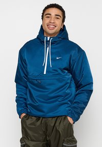 Blå nylon pullover-jakke med hætte, frontlynlås, elastiske manchetter og en brystlomme. Har et hvidt logo på øverste venstre side.