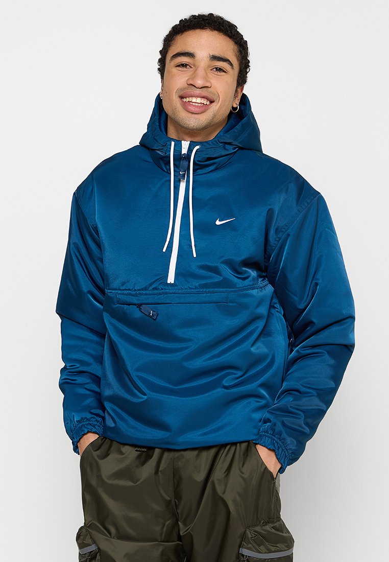 Blå nylon pullover-jakke med hætte, frontlynlås, elastiske manchetter og en brystlomme. Har et hvidt logo på øverste venstre side.