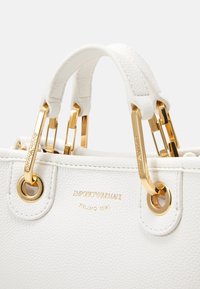 Borsa in pelle bianca con dettagli in metallo dorato, presenta doppi manici e logo impresso. Superficie strutturata e occhielli circolari visibili.