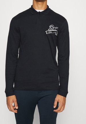 Man in een zwarte longsleeve poloshirt met een wit "Boss Athletic" logo op de borst en donkere broek, staande tegen een effen achtergrond.