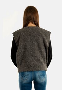 Gilet gris en feutre avec un col haut, sans manches, et texture visible. Porté par-dessus un haut à manches longues noir avec un jean en denim.