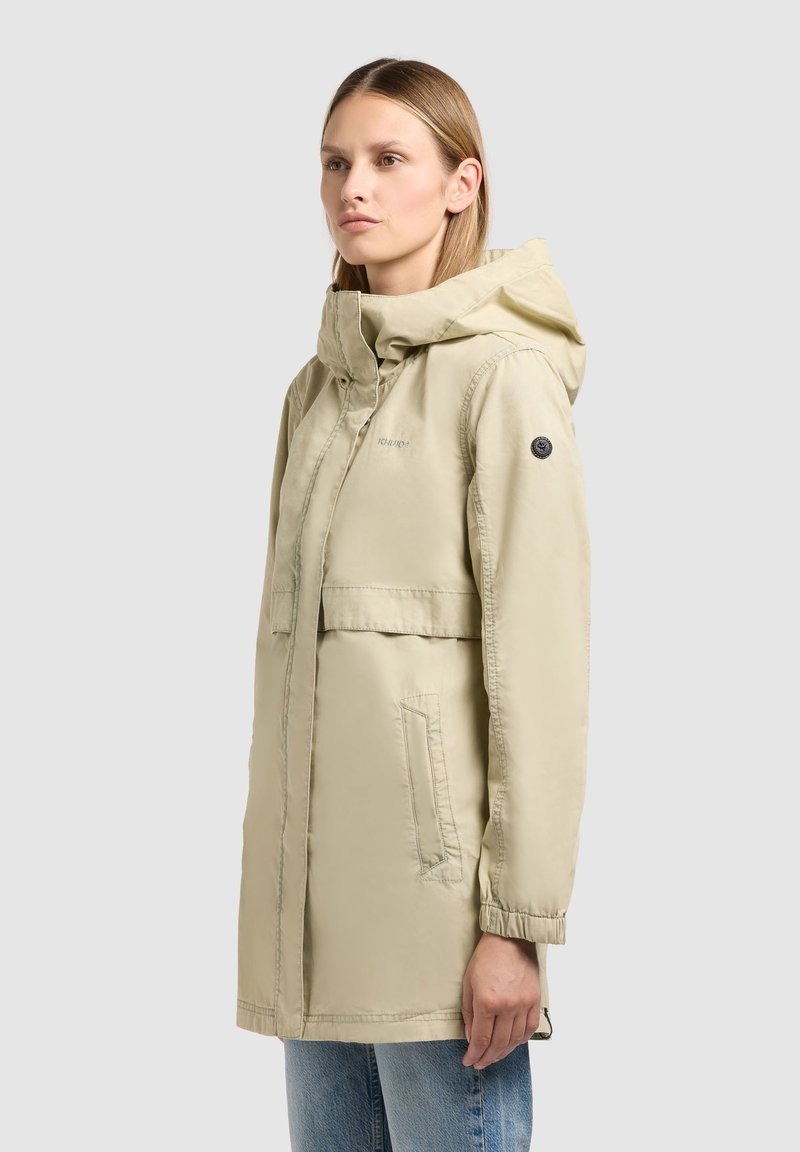Jeune femme portant une parka beige à capuche avec des poignets élastiques, debout de profil sur un fond clair uni.