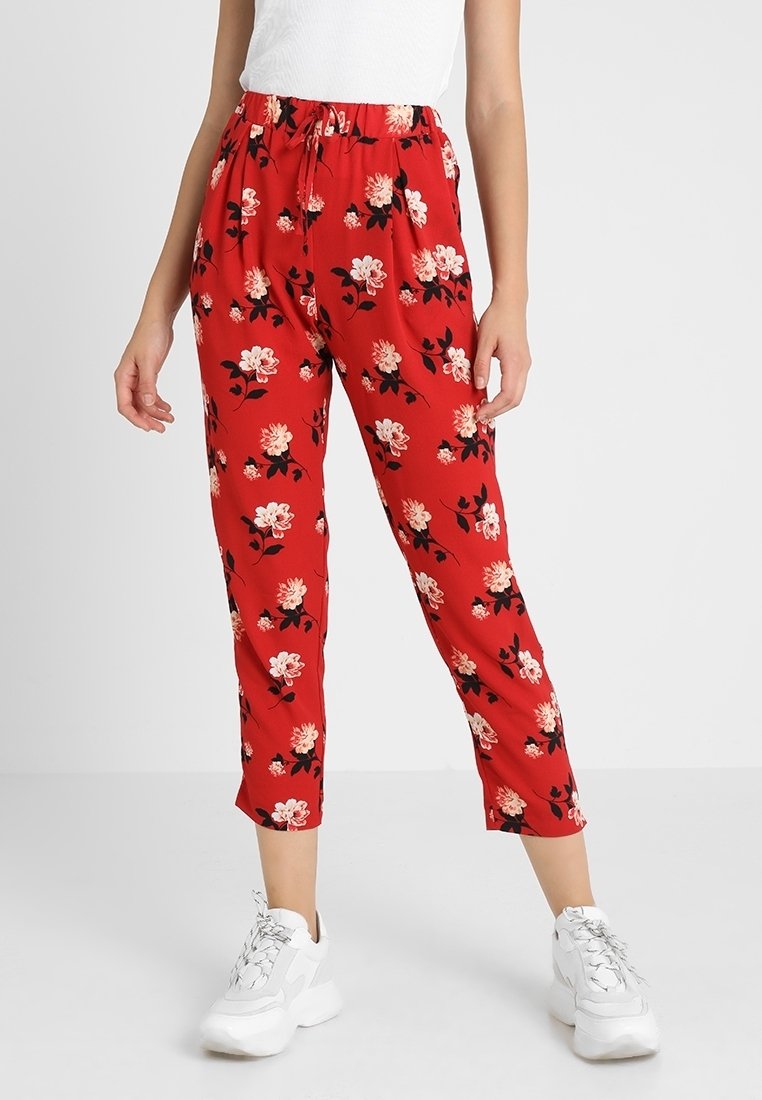 Femme portant un pantalon rouge court avec un motif floral blanc, associé à des baskets blanches et un haut blanc sans manches.