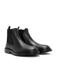 Bottes Chelsea en cuir noir avec panneaux latéraux élastiques, semelle plate et languettes pour faciliter l'enfilage. Texture lisse avec des détails minimalistes, idéales pour un usage décontracté.