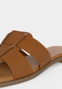 Καφέ δερμάτινο σανδάλι σλάιντ με φαρδιές διασταυρούμενες λουρίδες και επίπεδη σόλα σε απλό ανοιχτό φόντο.