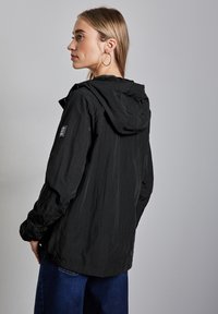 Svart lättviktsjacka med huva, i strukturerad tyg med vertikala ränder. Inkluderar en logotyp på vänster ärm. Bärs med vida blå jeans.