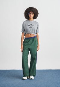 Γκρι κοντομάνικο φούτερ με εκτύπωση "Nike 1972", συνδυασμένο με σκούρο πράσινο παντελόνι φόρμας με μπλε ρίγες στο πλάι. Το μοντέλο φοράει λευκά αθλητικά παπούτσια.