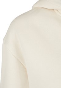 Gros plan d'un tissu couleur crème montrant la couture de l'épaule et de la capuche d'un vêtement avec une texture lisse et tricotée.