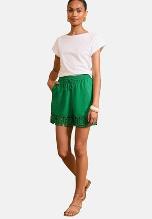 Grüne Baumwollshorts mit einem Kordelzugbund und Spitzenbesatz am Saum, kombiniert mit einem weißen Kurzarmoberteil und braunen Sandalen.