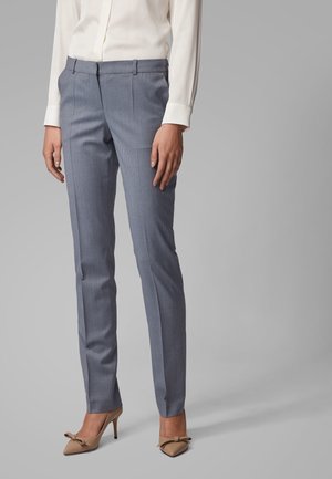 Femme portant un pantalon slim gris, des escarpins beige avec des nœuds et un chemisier blanc à manches longues sur un fond gris.