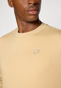 Beige genser laget av bomull, med rund hals og en subtil brodert logo i en lysere nyanse på brystet.