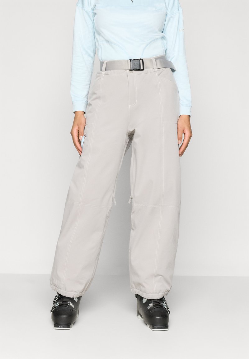 Calças de neve cinzentas com um cinto, apresentando bolsos com fechos de correr e costuras reforçadas, combinadas com botas de esqui pretas, usadas sobre uma camisa de manga longa azul clara.