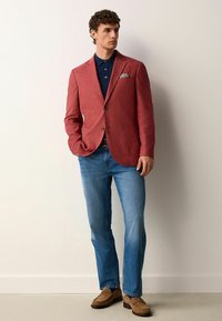 Roter Leinen-Blazer mit Reverskragen, frontalen Taschen und einem blauen Polo darunter. Kombiniert mit hellblauen Jeans und beigen Loafers.