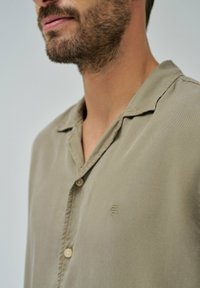 Camisa de manga corta color verde oliva con cuello, que presenta un cierre de botones y un sutil logo bordado en el lado izquierdo.