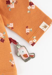 Baumwollbluse in Orange mit floralem Muster in Pink, Rot und Grün. Verfügt über ein Etikett mit einem Vogeldesign und einem strukturierten Schlaufenverschluss.