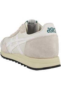 Zapatilla deportiva beige con materiales de malla y ante, que presenta un logo blanco de Asics, acentos texturizados y una suela de goma negra.