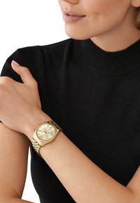 Orologio cronografo in oro con cinturino texturizzato, dotato di un quadrante rotondo, numerosi sottodial e una finitura lucida, indossato su un polso.