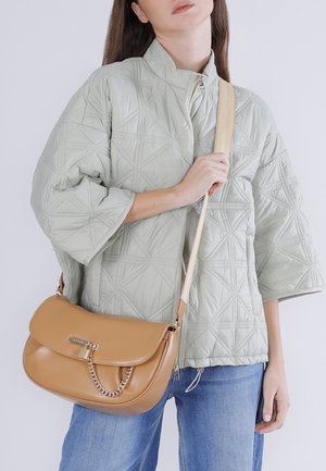 Vrouw met een lichtgrijze doorgestikte jas en een blauwe spijkerbroek, die een beige leren schoudertas met gouden kettingdetail vasthoudt.