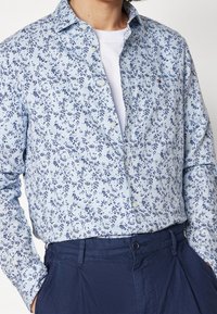 Světle modrá košile na knoflíky s květinovým vzorem v námořnické modři. Košile má klasický límec a manžety na jedno knoflíček, doplněné tmavými kalhotami.