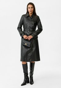 Zwarte leren trenchcoat met dubbele knopen, een ceintuur in de taille en lange mouwen. Gecombineerd met zwarte laarzen tot de knie en een kleine handtas.