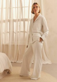 Femme se tenant dans une pièce doucement éclairée, v�êtue d'un ensemble de pyjama beige clair et ample, les mains dans les poches, avec des rideaux transparents et un lit en arrière-plan.