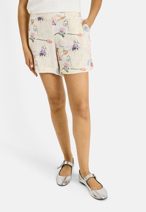 Femme portant un short couleur crème avec des motifs floraux et de l'art linéaire, chaussures Mary Jane argentées, debout avec une main dans la poche sur fond blanc.