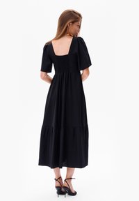 Schwarzes Midi-Kleid mit kurzen Puffärmel, quadratischem Rückenteil, gerafftem Oberteil und gestuftem Rüschensaum. Kombiniert mit schwarzen hochhackigen Sandalen.