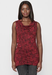 Débardeur rouge sans manches avec un col drapé, arborant un motif de feuilles noires. Prenu sur fond blanc uni, porté par un modèle.