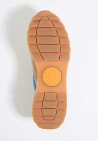 Suela de goma en tono marrón con tracción estampada. Presenta un logo circular en naranja en el centro. La sección superior muestra ante azul y beige.
