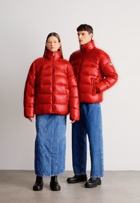 Κόκκινα μπουφάν puffer με γυαλιστερό φινίρισμα, με ψηλό γιακά και κλείσιμο με φερμουάρ. Τα μοντέλα φορούν μπλε τζιν παντελόνια και μαύρες μπότες.