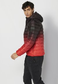 Giacca imbottita con una gradazione che va dal nero scuro al rosso brillante, dotata di cappuccio e tasche con cerniera. Superficie con texture e trapuntatura orizzontale.