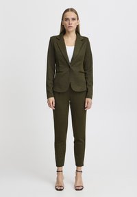 Olivengrüner maßgeschneiderter Anzug aus strukturiertem Stoff, slim-fit Blazer und schmal geschnittene Hose. Kombiniert mit schwarzen Riemchen-Pumps.