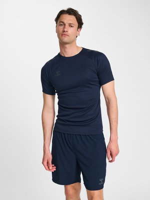 Mężczyzna ubrany w granatową sportową koszulkę i krótkie spodenki stoi na jednolitym białym tle.