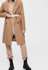 Kabát z camelové vlny se směsí, se zapínáním na dva knoflíky, velkými klopami a bočními kapsami, stažený v pase bílým páskem. Nositelný přes béžové tepláky a černé boty.