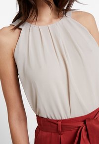 Lys beige ærmeløs bluse med en sammensamlet halsudskæring, der har et blødt stof. Kombineret med et rødt taljebånd som accent.