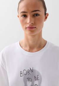 Witte katoenen t-shirt met een ronde hals en korte mouwen, voorzien van een grafisch ontwerp met de tekst "BORN TO BE WILD" in donkere letters.