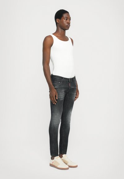 Jeans skinny negros con detalles desgastados, combinados con una camiseta sin mangas blanca y zapatillas beige con acentos naranjas. Diseño simple, estilo ajustado.