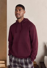 Burgundy huva sweatshirt med en framlappficka, bärs med mönstrade rutiga pyjamasbyxor, med en mjuk textur och avslappnad design.