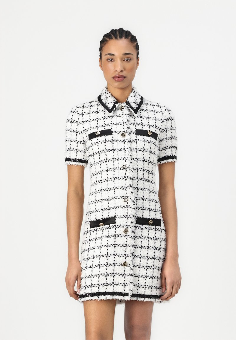 White and black tweed dress with short sleeves, collar, front pockets, gold buttons, and contrasting trim detailing. Textured fabric with a classic pattern.