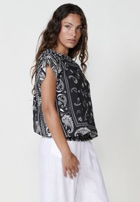 Haut à motif paisley noir et blanc avec des manches courtes, coupe décontractée et ourlet froncé, associé à un pantalon large de couleur claire.
