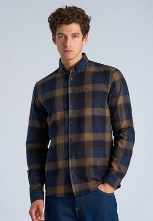 Mann mit lockigem Haar, der ein marineblau-braunes kariertes Hemd mit Knöpfen trägt und mit einer Hand in der Tasche seiner blauen Jeans nach vorne schaut.