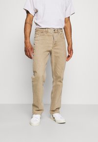 Jeans de mezclilla beige con corte recto, diseño de cinco bolsillos y costuras visibles; combinados con zapatillas blancas y una camiseta clara.