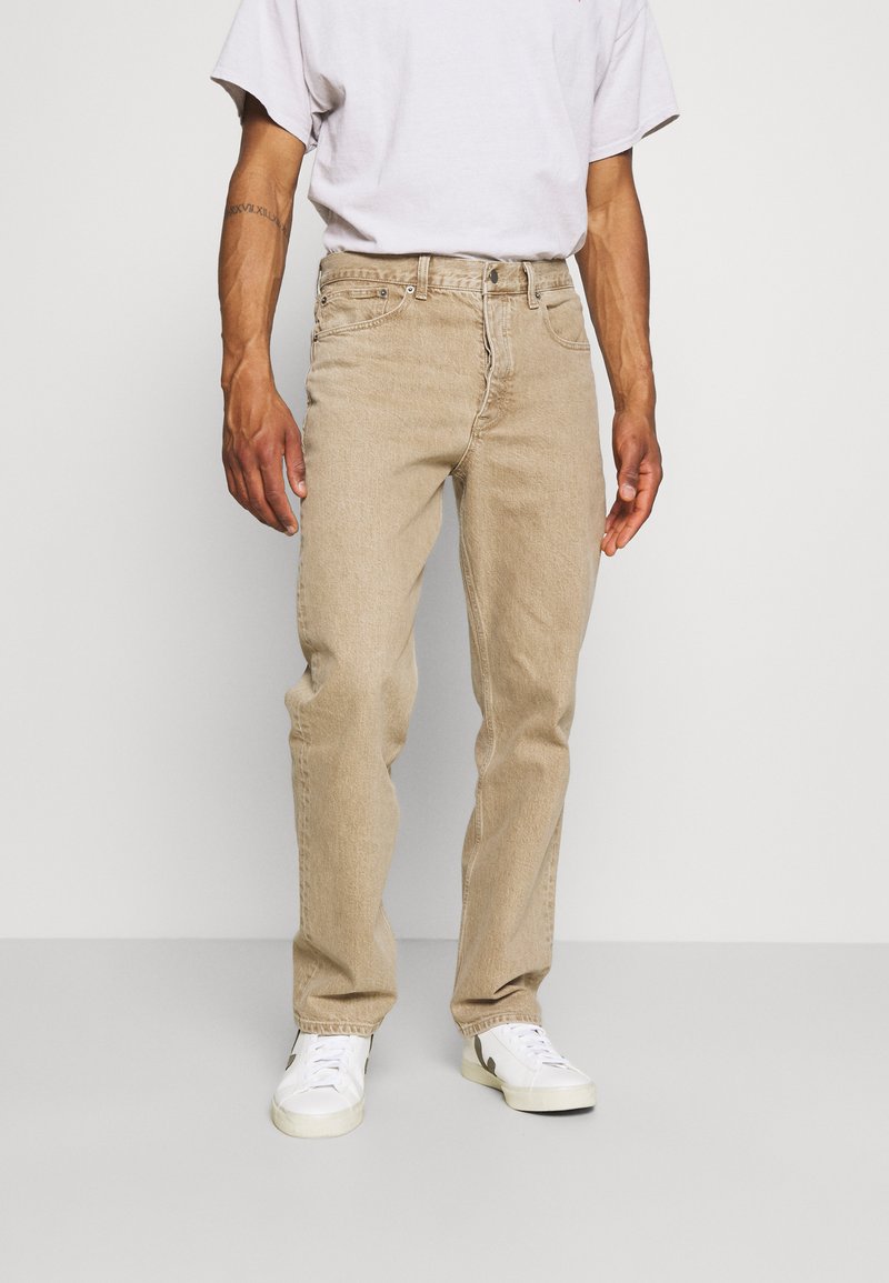 Jeans de mezclilla beige con corte recto, diseño de cinco bolsillos y costuras visibles; combinados con zapatillas blancas y una camiseta clara.